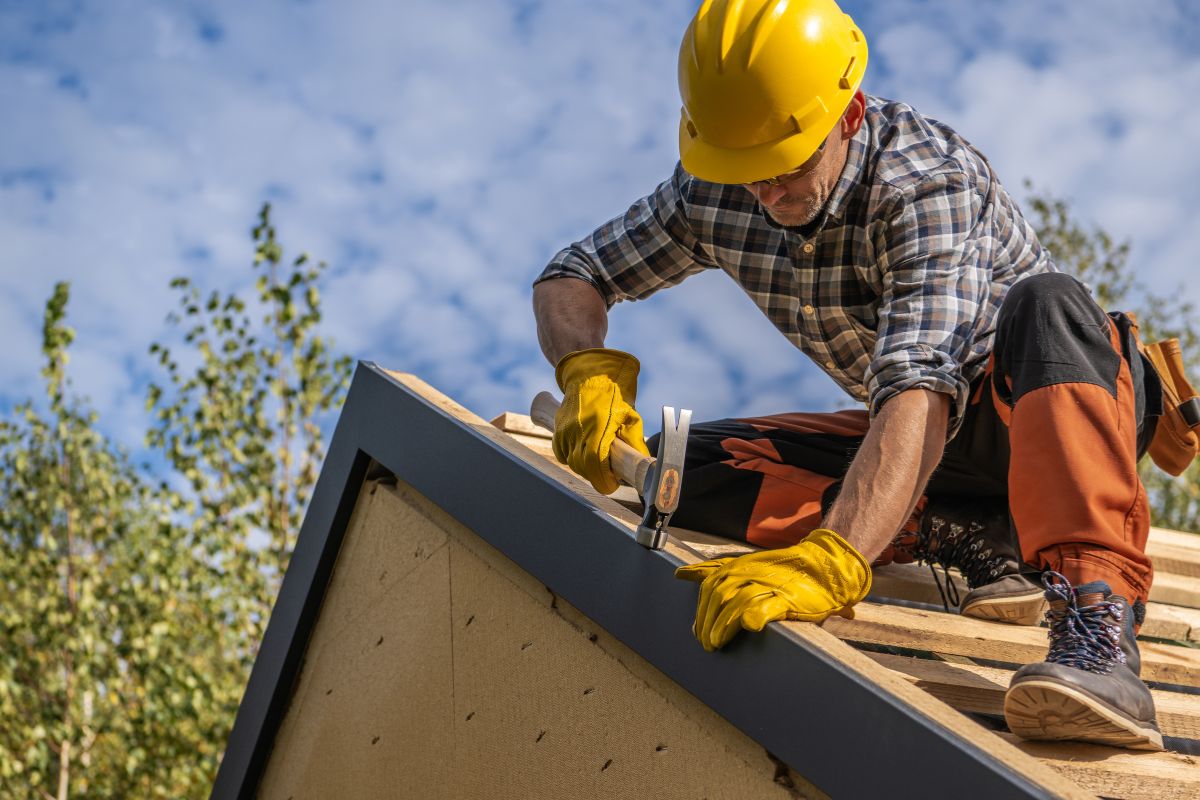 A construction worker in a hard hat and yellow safety vest is actively working on a roof, focusing on his task.