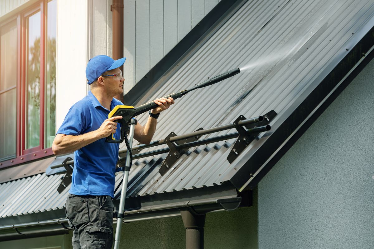 A man wearing a blue shirt and hat is cleaning a roof with a brush, standing on a ladder.
