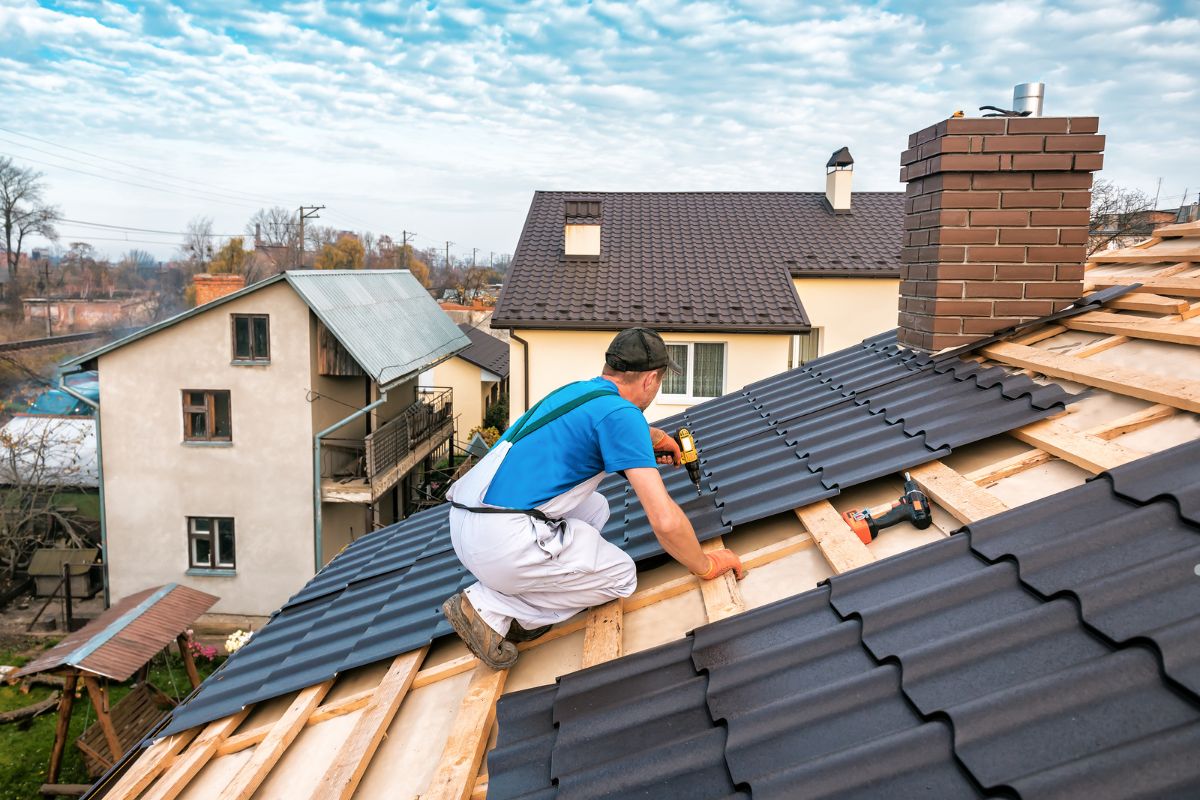 A man is installing a metal roof, standing on a sloped surface with tools in hand, focused on his work.