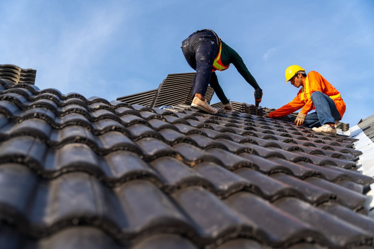 Two men are installing roofing materials under a clear blue sky.